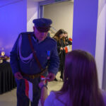 Na obrazie starszy mężczyzna w mundurze wręcza flagę dziewczynce. Po lewej stół z jedzeniem, tło fioletowe. W tle inni ludzie z flagami.