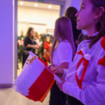 Grupa osób zebrana w jasnym pomieszczeniu trzyma biało-czerwone flagi. Ubrani w białe koszule z czerwonymi akcentami. Światło tworzy purpurowy ton.