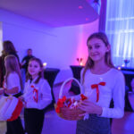 Na zdjęciu widać trzy dziewczynki w białych bluzkach z czerwonymi wstążkami, trzymają koszyki. Tło jest jasno oświetlone na niebiesko i różowo. W tle widać ludzi oraz zasłony okienne.