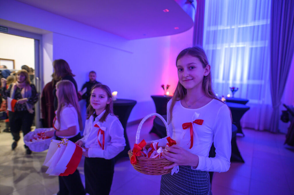 Na zdjęciu kilka dziewczynek w białych bluzkach z czerwonymi wstążkami, trzymają koszyki i flagi Polski. Tło w fioletowym świetle, ciemne stoły, zasłony. W tle widoczni ludzie wchodzący do pomieszczenia.