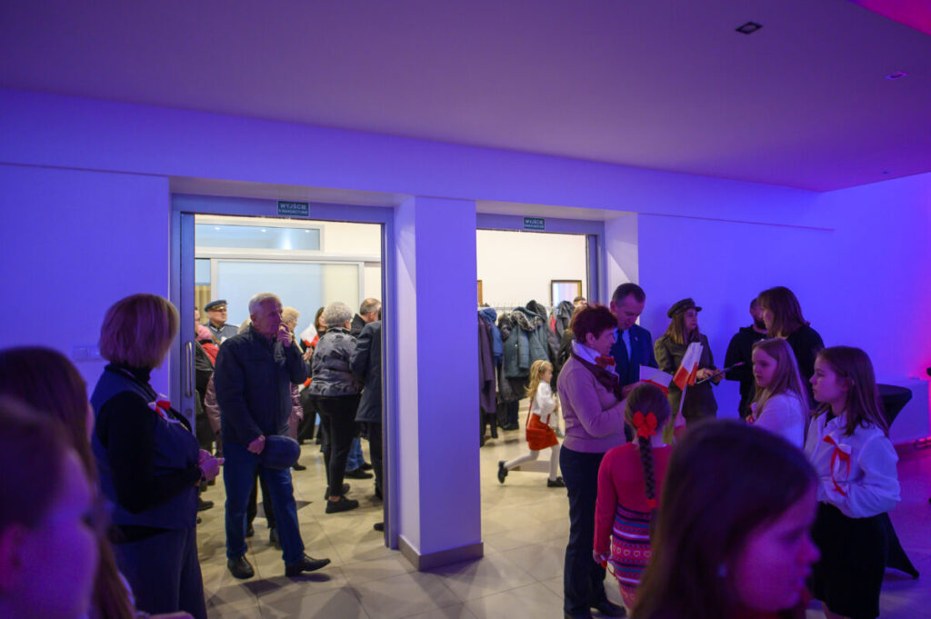 Na zdjęciu widać grupę ludzi w jasnym pomieszczeniu. W tle płaszcze, a ściany są oświetlone na niebiesko i fioletowo. Niektórzy trzymają polskie flagi.