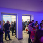 Na zdjęciu widać grupę ludzi w jasnym pomieszczeniu. W tle płaszcze, a ściany są oświetlone na niebiesko i fioletowo. Niektórzy trzymają polskie flagi.