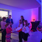 Grupa ludzi stoi w jasnym pomieszczeniu. Młode dziewczyny trzymają flagi i koszyk. Tło jest niebieskie z różowym światłem.