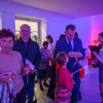 Grupa ludzi w sali z niebieskim oświetleniem trzyma polskie flagi. Mężczyzna w garniturze rozmawia z dziewczynką w czerwieni. W tle widać kwiaty na stolikach.