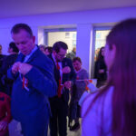 Grupa ludzi w jasnym pomieszczeniu ozdabia siebie biało-czerwonymi wstążkami. Fioletowe oświetlenie dodaje nastroju. W tle inne osoby i stół z jedzeniem.