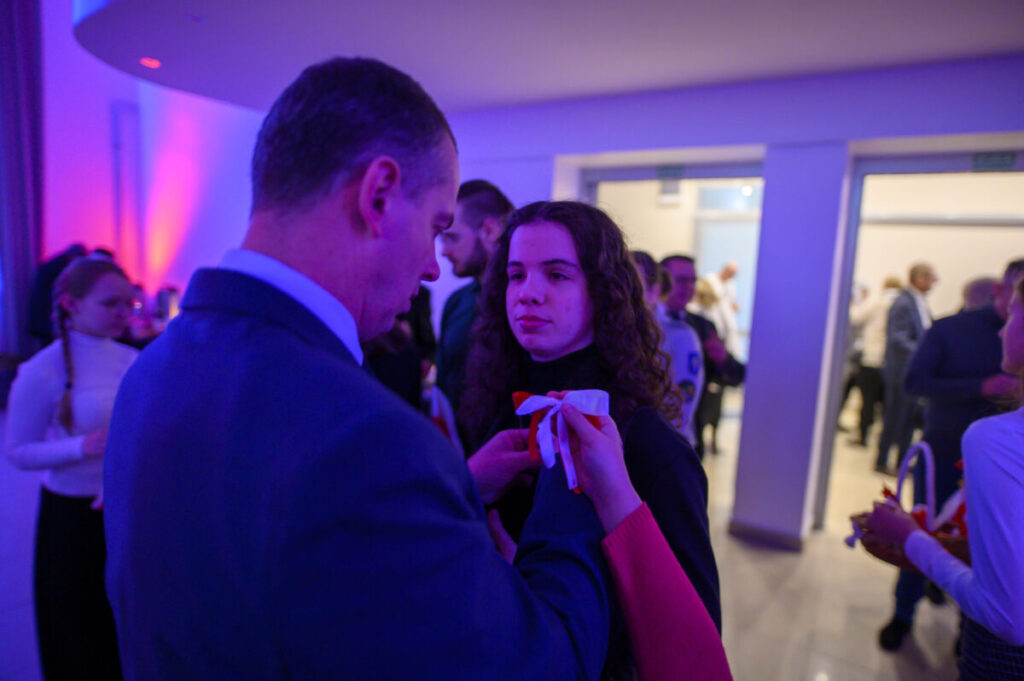 Na zdjęciu widać mężczyznę przypinającego kotylion kobiecie. Dominują odcienie niebieskiego i fioletowego. Tło to eleganckie wnętrze z ludźmi.