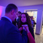 Na zdjęciu widać mężczyznę przypinającego kotylion kobiecie. Dominują odcienie niebieskiego i fioletowego. Tło to eleganckie wnętrze z ludźmi.