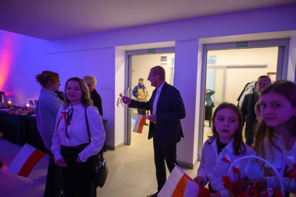 Grupa osób w eleganckich strojach, trzyma biało-czerwone flagi. Tło oświetlone na fioletowo. W tle stół z przekąskami.