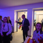 Grupa osób w eleganckich strojach, trzyma biało-czerwone flagi. Tło oświetlone na fioletowo. W tle stół z przekąskami.