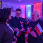 Trzy osoby rozmawiają w sali z niebiesko-czerwonym oświetleniem. Dwie z nich w czarnych sutannach trzymają polskie flagi. W tle widać ludzi i zasłony.