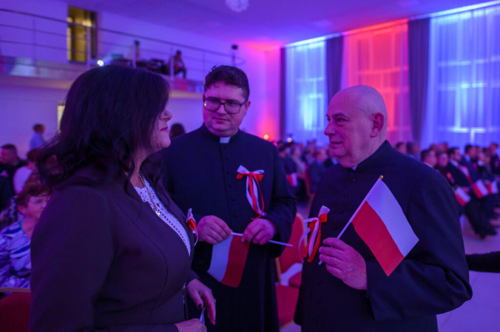 Trzy osoby rozmawiają w oświetlonej sali z niebieskim i czerwonym światłem. Dwie osoby w czarnych strojach trzymają polskie flagi. Tło to zebrany tłum siedzący na czerwonych krzesłach.