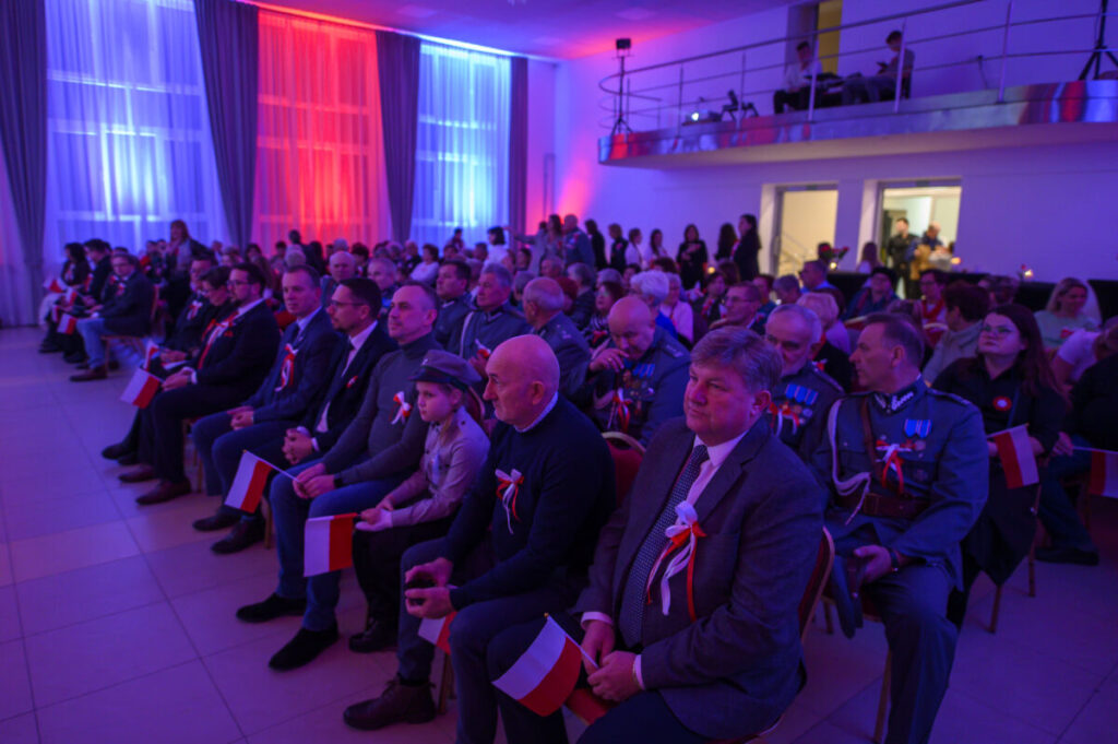 Na zdjęciu widzimy grupę ludzi na uroczystości z czerwono-niebieskim oświetleniem. Siedzą z polskimi flagami w dłoniach. W tle są ozdobne zasłony i podwyższenie.