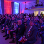 Na zdjęciu widzimy grupę ludzi na uroczystości z czerwono-niebieskim oświetleniem. Siedzą z polskimi flagami w dłoniach. W tle są ozdobne zasłony i podwyższenie.