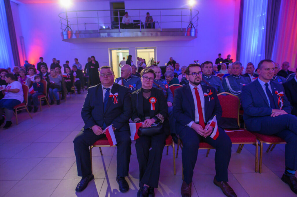 Grupa ludzi w eleganckich strojach siedzi w sali oświetlonej na niebiesko i różowo. Trzymają flagi Polski. Tło to balkon z osobami obserwującymi.