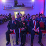 Grupa ludzi w eleganckich strojach siedzi w sali oświetlonej na niebiesko i różowo. Trzymają flagi Polski. Tło to balkon z osobami obserwującymi.