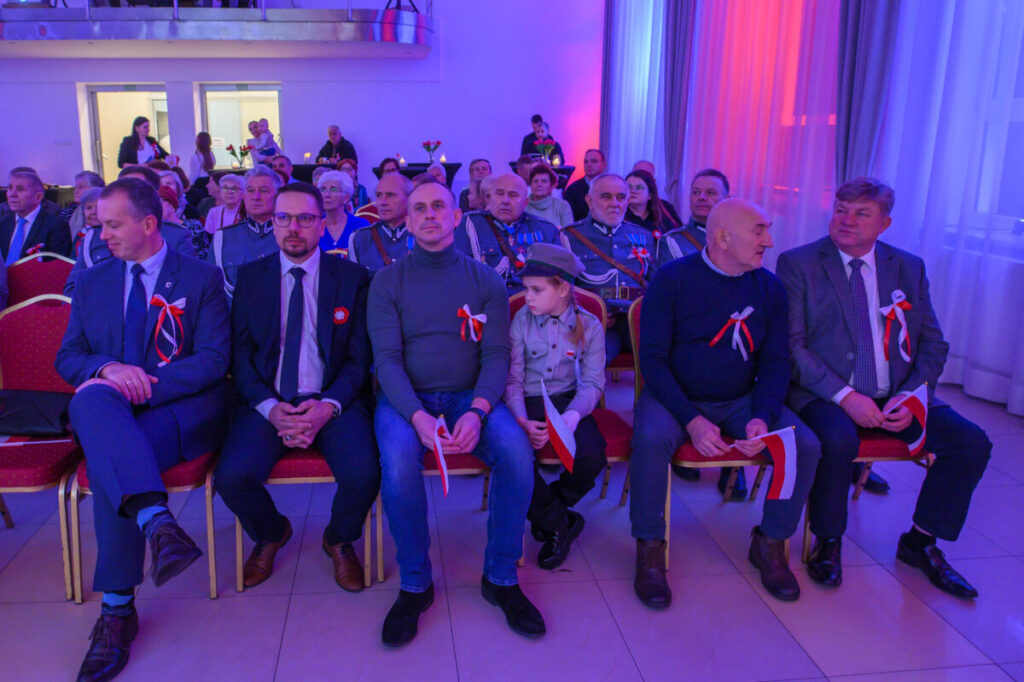 Grupa ludzi siedzi w formalnej sali, ubrana elegancko z biało-czerwonymi kotylionami. Ściany oświetlone są na niebiesko i czerwono. Niektórzy trzymają polskie flagi.