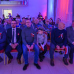 Grupa ludzi siedzi w formalnej sali, ubrana elegancko z biało-czerwonymi kotylionami. Ściany oświetlone są na niebiesko i czerwono. Niektórzy trzymają polskie flagi.