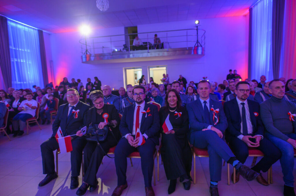 Grupa ludzi siedzi w sali oświetlonej na niebiesko i różowo. Mają przypięte kokardy, niektórzy trzymają flagi Polski. Tło jest jasne, z balkonem i publicznością.