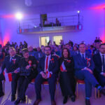 Grupa ludzi siedzi w sali oświetlonej na niebiesko i różowo. Mają przypięte kokardy, niektórzy trzymają flagi Polski. Tło jest jasne, z balkonem i publicznością.