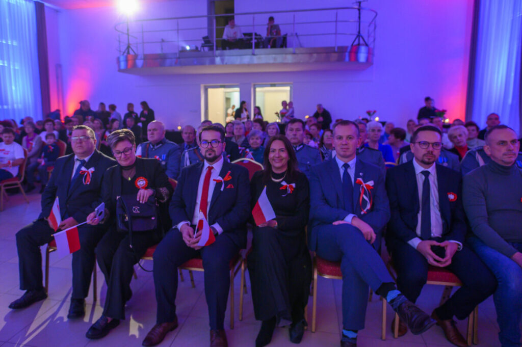 Grupa ludzi siedzi w kilku rzędach na uroczystości. Mają biało-czerwone kokardy i flagi Polski. Tło oświetlone na niebiesko i różowo.