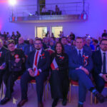 Grupa ludzi siedzi w kilku rzędach na uroczystości. Mają biało-czerwone kokardy i flagi Polski. Tło oświetlone na niebiesko i różowo.