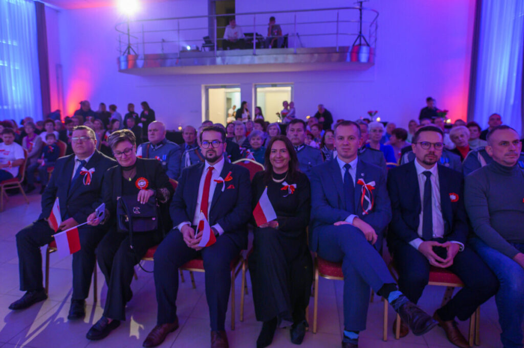 Grupa ludzi siedzących na uroczystości, ubrana elegancko, część z flagami Polski. Tło z niebieskim i fioletowym oświetleniem. Sala wypełniona osobami na krzesłach.