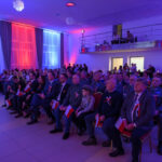 Na zdjęciu grupa ludzi siedzi w sali z niebiesko-czerwonym oświetleniem. Wszyscy mają biało-czerwone wstążki i flagi. Tło jest jasne, z ozdobnymi zasłonami.