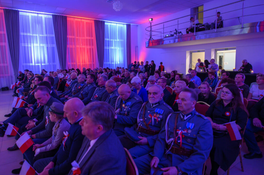 Sala pełna ludzi w mundurach i formalnych strojach. Niebiesko-czerwone światła oświetlają scenę. Uczestnicy trzymają polskie flagi.