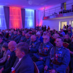 Sala pełna ludzi w mundurach i formalnych strojach. Niebiesko-czerwone światła oświetlają scenę. Uczestnicy trzymają polskie flagi.