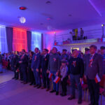 Grupa osób w eleganckich strojach stoi w dużej sali. Dominuje oświetlenie w kolorach niebieskim i czerwonym. Uczestnicy mają przypięte biało-czerwone kokardy.