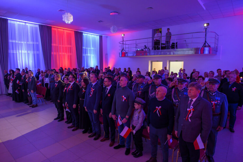 Grupa osób stoi na uroczystości, ubrana w patriotyczne stroje. Tło podświetlone jest na czerwono, biało i niebiesko. Osoby trzymają małe flagi Polski.