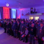 Grupa osób stoi na uroczystości, ubrana w patriotyczne stroje. Tło podświetlone jest na czerwono, biało i niebiesko. Osoby trzymają małe flagi Polski.