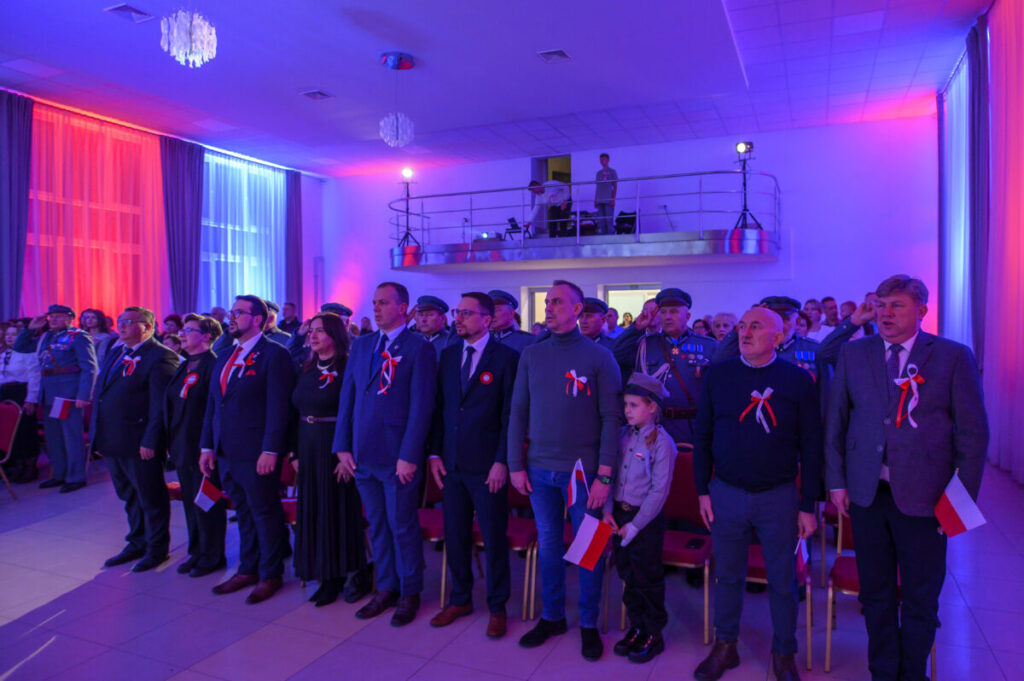 Grupa ludzi stoi w sali oświetlonej na czerwono-niebiesko. Uczestnicy mają biało-czerwone ozdoby i trzymają polskie flagi. W tle balkon z obserwatorami.