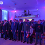 Grupa ludzi stoi w sali oświetlonej na czerwono-niebiesko. Uczestnicy mają biało-czerwone ozdoby i trzymają polskie flagi. W tle balkon z obserwatorami.