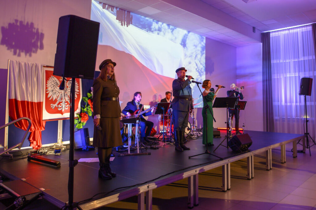 Na zdjęciu jest grupa muzyków na scenie, otoczeni flagą Polski. Tło wypełnia biało-czerwona flaga i projekcja. Muzycy noszą eleganckie stroje, scena jest oświetlona na fioletowo.