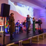 Na zdjęciu jest grupa muzyków na scenie, otoczeni flagą Polski. Tło wypełnia biało-czerwona flaga i projekcja. Muzycy noszą eleganckie stroje, scena jest oświetlona na fioletowo.