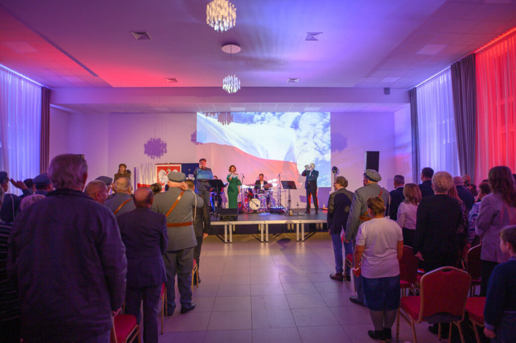 Na zdjęciu jest koncert z widownią, na scenie zespół. Światła w kolorach czerwono-białych, w tle projektor z flagą. Sala jest przestronna z krzesłami.