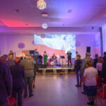 Na zdjęciu jest koncert z widownią, na scenie zespół. Światła w kolorach czerwono-białych, w tle projektor z flagą. Sala jest przestronna z krzesłami.