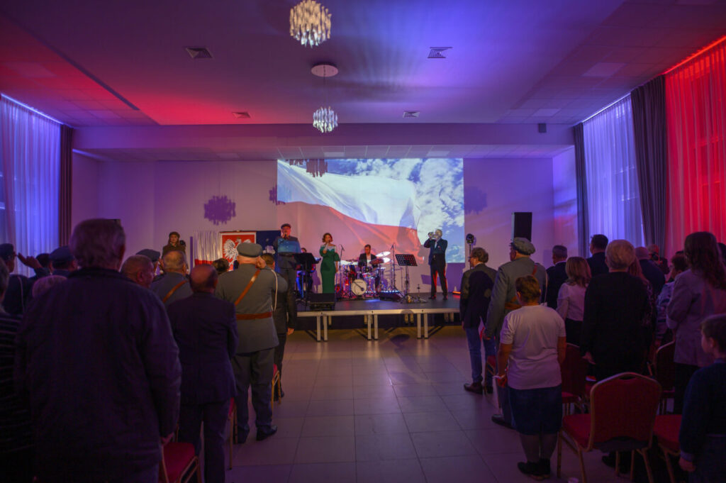 Koncert w sali, czerwone i niebieskie światła, polska flaga w tle. Orkiestra gra przed zgromadzoną publicznością. Dekoracje w stylu patriotycznym.