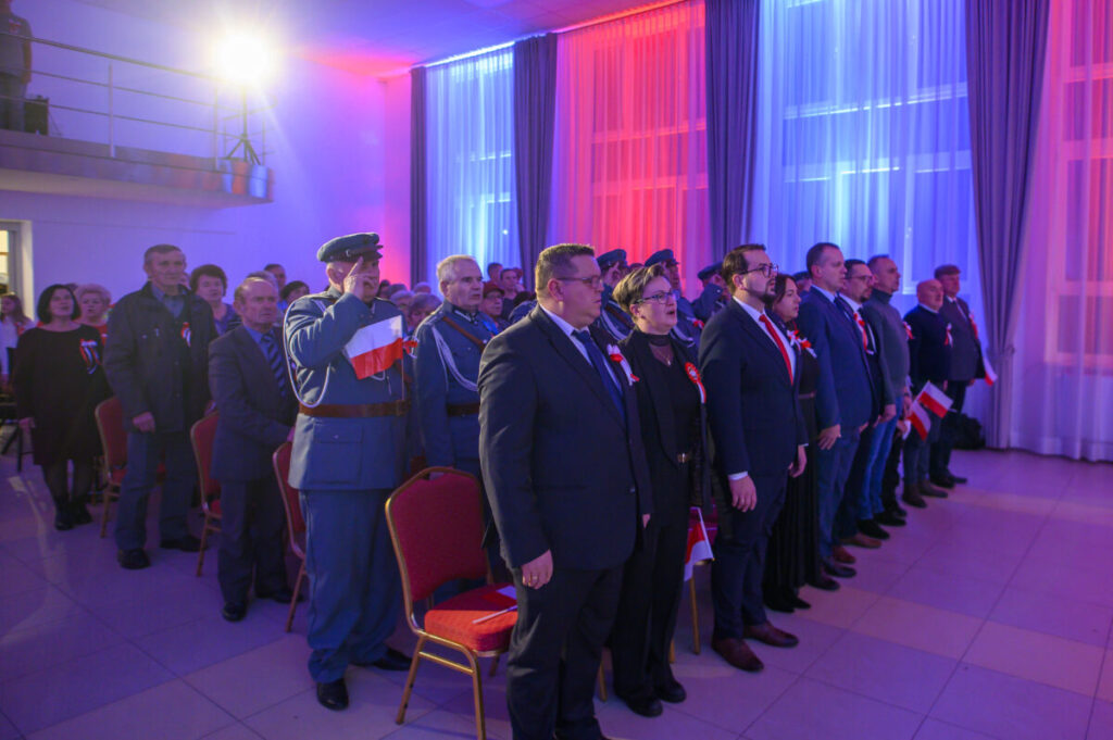 Grupa ludzi stoi w eleganckiej sali, ubrani w formalne stroje, z biało-czerwonymi flagami. Tło oświetlone jest na niebiesko i czerwono. Wydarzenie ma oficjalny charakter.