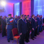 Grupa ludzi stoi w eleganckiej sali, ubrani w formalne stroje, z biało-czerwonymi flagami. Tło oświetlone jest na niebiesko i czerwono. Wydarzenie ma oficjalny charakter.