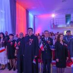 Grupa ludzi ubrana odświętnie z polskimi flagami i kotylionami. Tło oświetlone w kolorach biało-czerwonych i niebieskich. Sala z wysokim sufitem i balkonem.