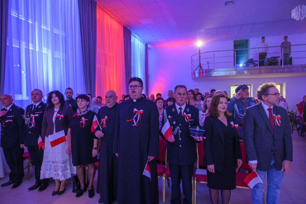 Grupa ludzi w oficjalnych strojach z biało-czerwonymi wstążkami. Sala udekorowana światłami w kolorach flagi Polski. Tło z białymi i czerwonymi zasłonami.