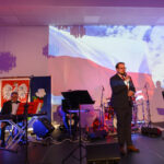 Na scenie mężczyzna przemawia, w tle wyświetlona flaga Polski. Obok ustawione są instrumenty muzyczne i sprzęt nagłaśniający. Oświetlenie w ciepłych kolorach.