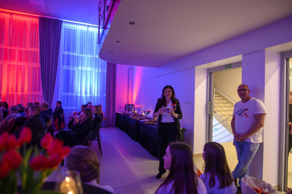 Spotkanie w sali z niebiesko-czerwonym oświetleniem. Ludzie siedzą na krzesłach, w tle stół z jedzeniem. Dwie osoby stoją przy drzwiach.