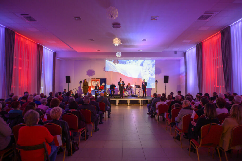 Widok na salę koncertową z ludźmi siedzącymi na czerwonych krzesłach. Scena oświetlona na niebiesko i czerwono, widoczny zespół muzyczny. W tle polska flaga na ekranie.