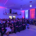 Sala pełna ludzi siedzących na uroczystości, z kolorowym oświetleniem w niebieskich i czerwonych tonach. W tle widoczne są zasłony i scena z oświetleniem. Uczestnicy trzymają polskie flagi.