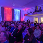 Grupa ludzi siedzi w sali oświetlonej na czerwono, niebiesko i biało. Tło stanowią zasłony w ciemnych odcieniach. Na piętrze są widoczni technicy przy sprzęcie.