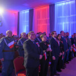 Uroczystość w sali z postaciami w oficjalnych strojach, tło w kolorach niebieskim i czerwonym, flagi biało-czerwone.