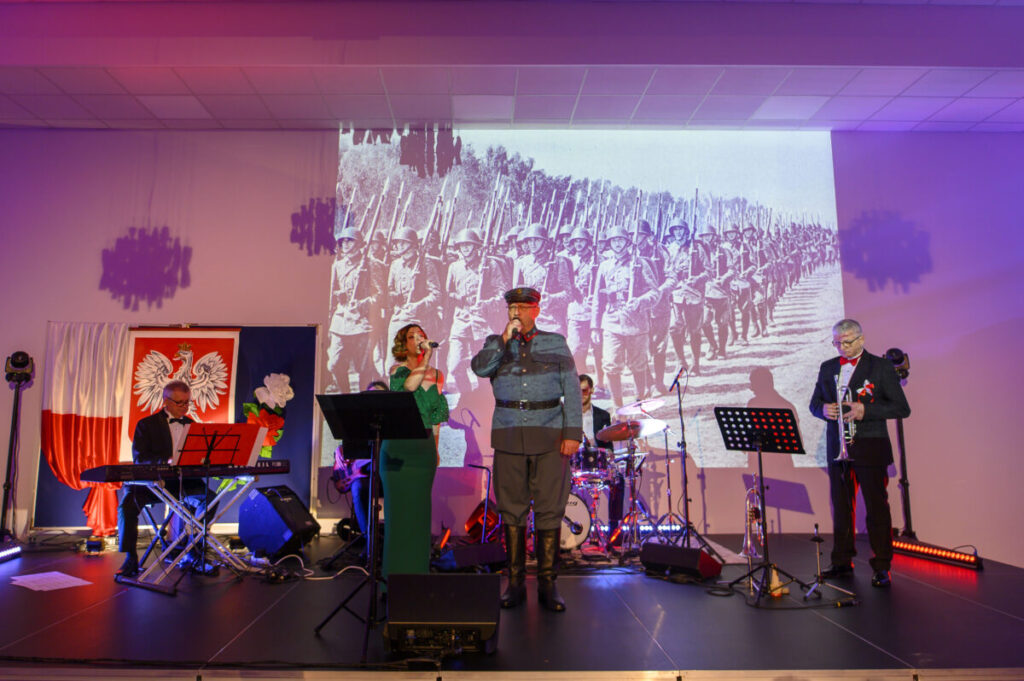 Grupa muzyków na scenie, w tle czarno-białe zdjęcie żołnierzy. Dominują kolory czerwony i biały z flagą Polski. Sala oświetlona fioletowym światłem.